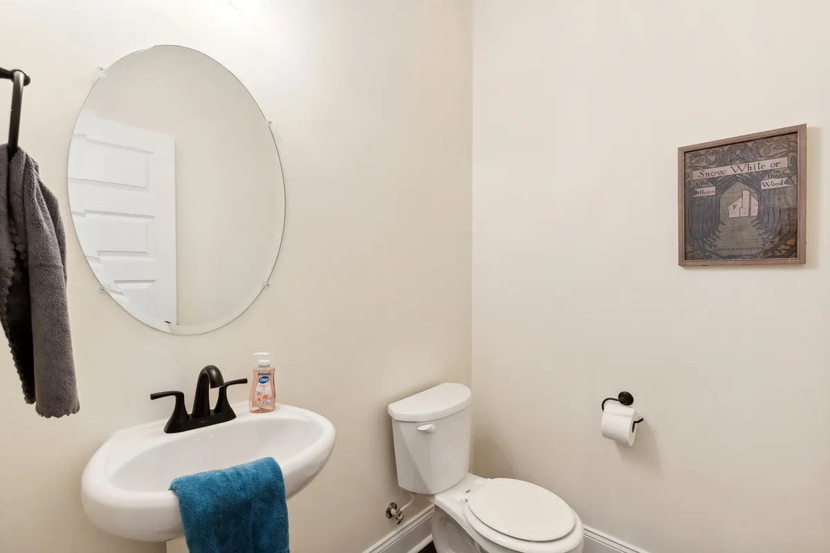 Depicts a fresh and welcoming half-bathroom with a round mirror, sink, toilet, framed wall art, and towel rack. The setting is clean, well-lit, and easily accessible—ideal for quick bathroom stops during meal prep or gatherings.