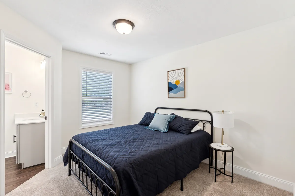 This serene bedroom features a bed dressed in navy blue bedding, a nightstand with a lamp, a window with blinds, and calming wall art. The layout encourages both restful sleep and quiet moments for reading or working, with ample furnishings for comfort and storage.