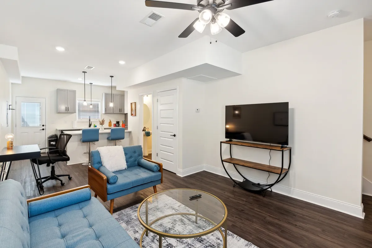 A contemporary, open-plan space featuring a blue couch, glass coffee table, TV, and a dining area with bar stools visible in the background. The scene is bathed in natural light, and the space feels modern and connected.