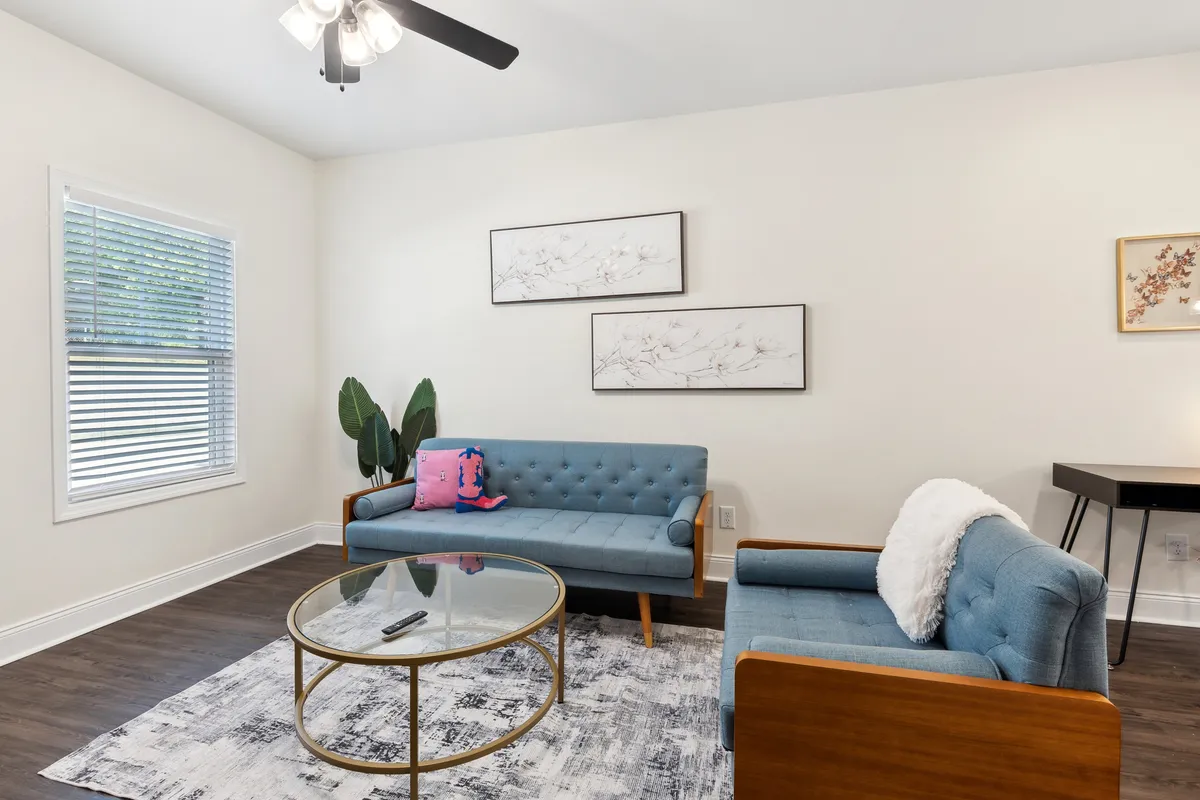 A bright, cozy living area with blue tufted couches arranged for conversation, a round glass coffee table, decorative wall art, and an indoor plant. Natural light streams through the room, highlighting the plush furnishings and sociable setup.