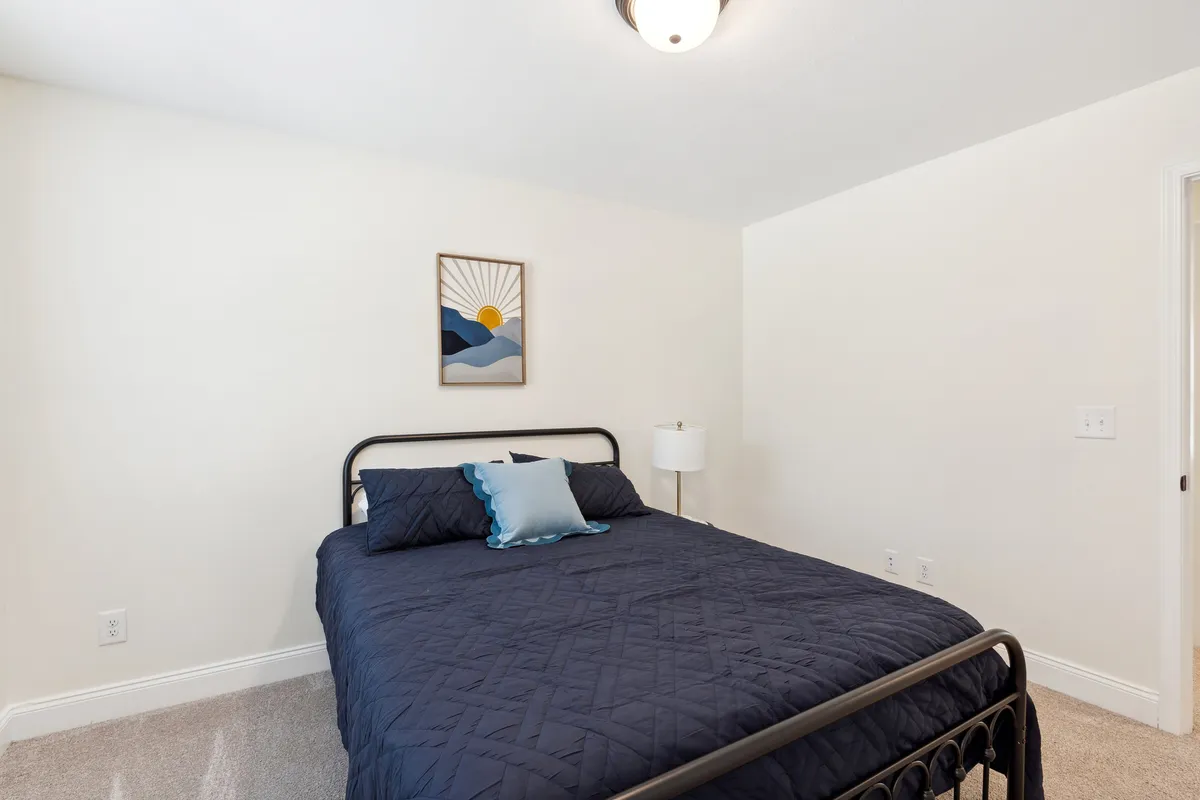 A tranquil bedroom scene featuring a bed with navy blue bedding, decorative pillows, a lamp, and framed artwork. The space is bathed in gentle daylight, reinforcing a sense of coziness and comfort.